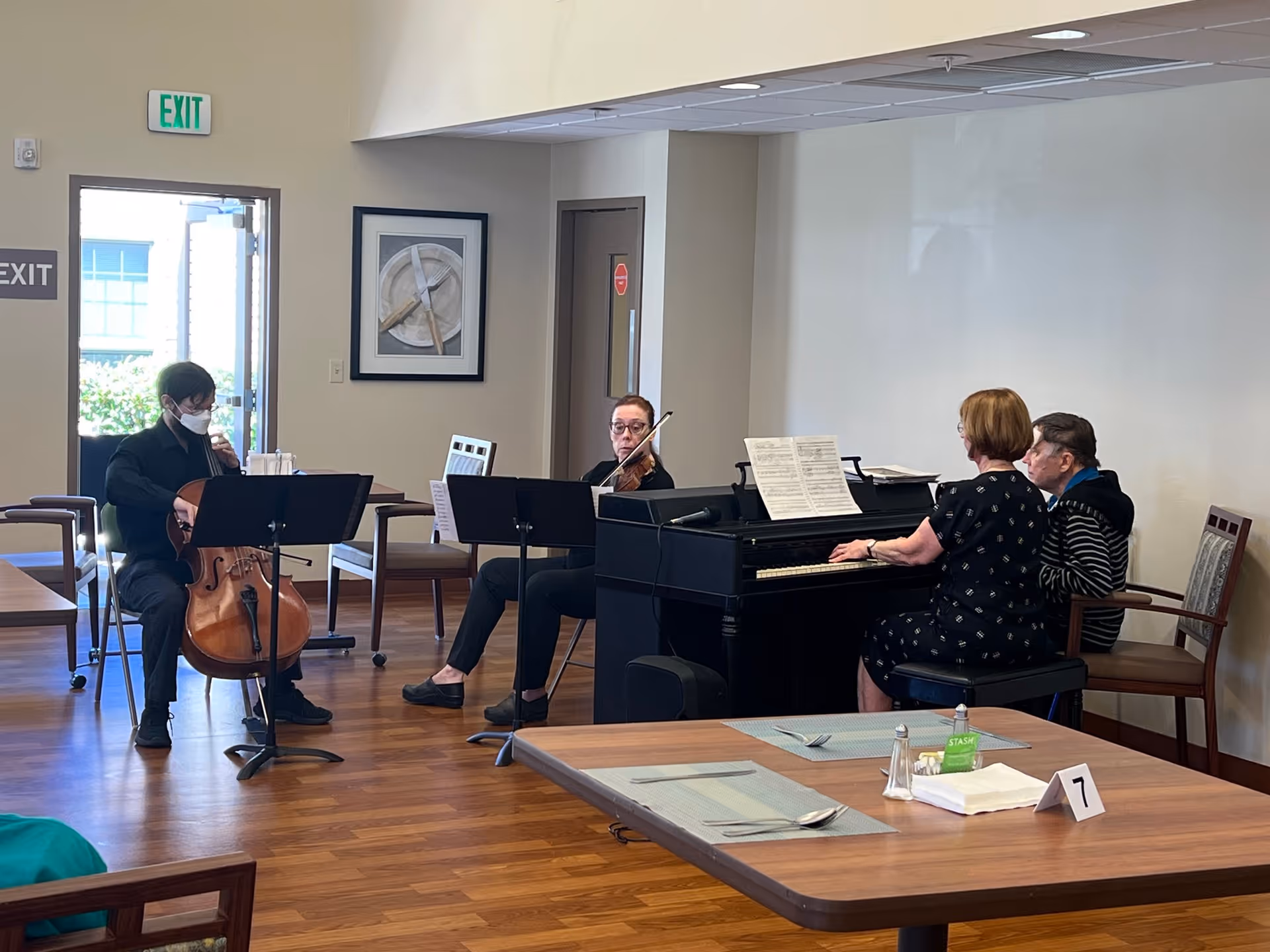 A small musical performance in a senior living facility common area. A cellist wearing a face mask and a violinist are playing music, while a woman plays the piano. An elderly person sits next to the pianist, listening. The room has wooden floors, tables with placemats and utensils, and an exit door in the background.