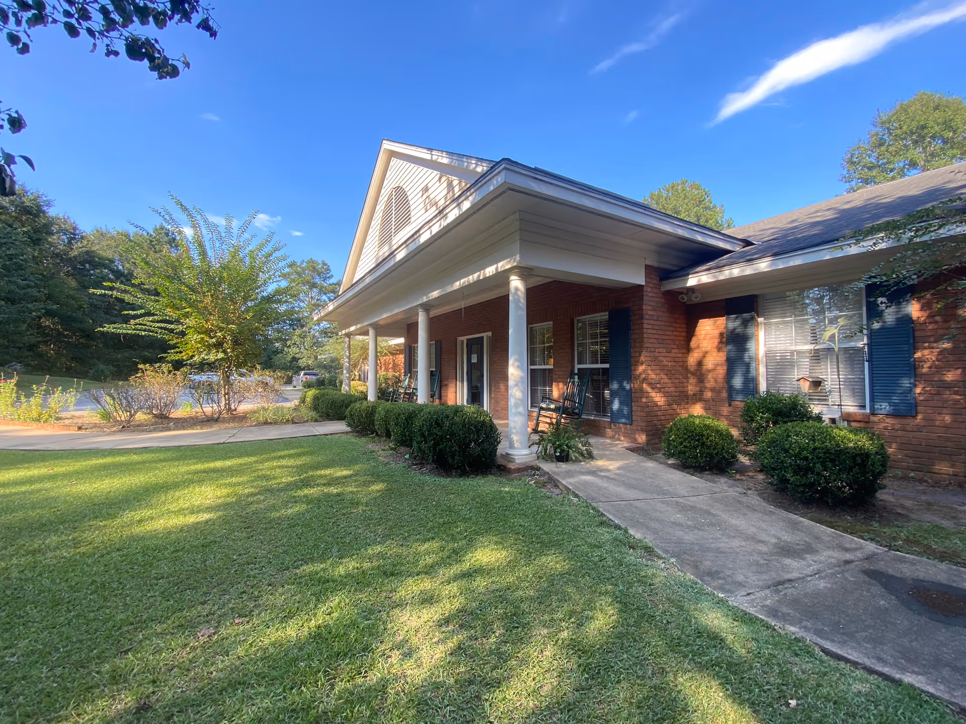 Front exterior of a single-story brick building with a covered porch, white columns, rocking chairs, and a grassy lawn.