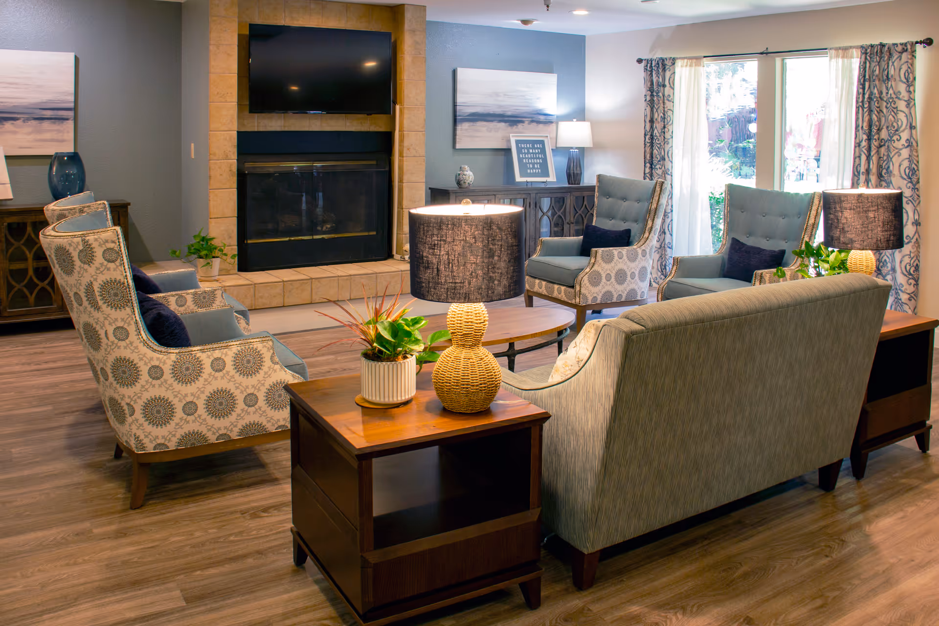 Cozy senior living lounge with patterned chairs, a central sofa, fireplace, TV, and lamps.