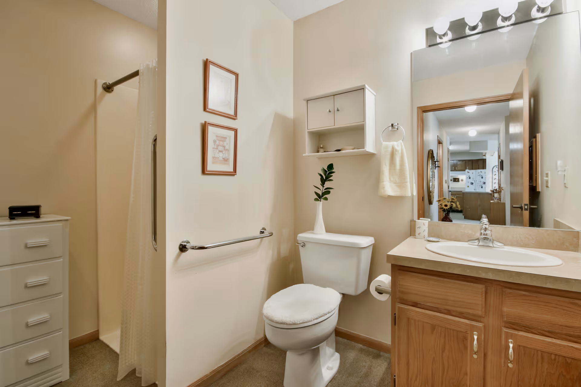 Well-lit bathroom with a toilet, sink vanity and mirror, a grab bar, shelving, and a walk-in shower with a curtain.