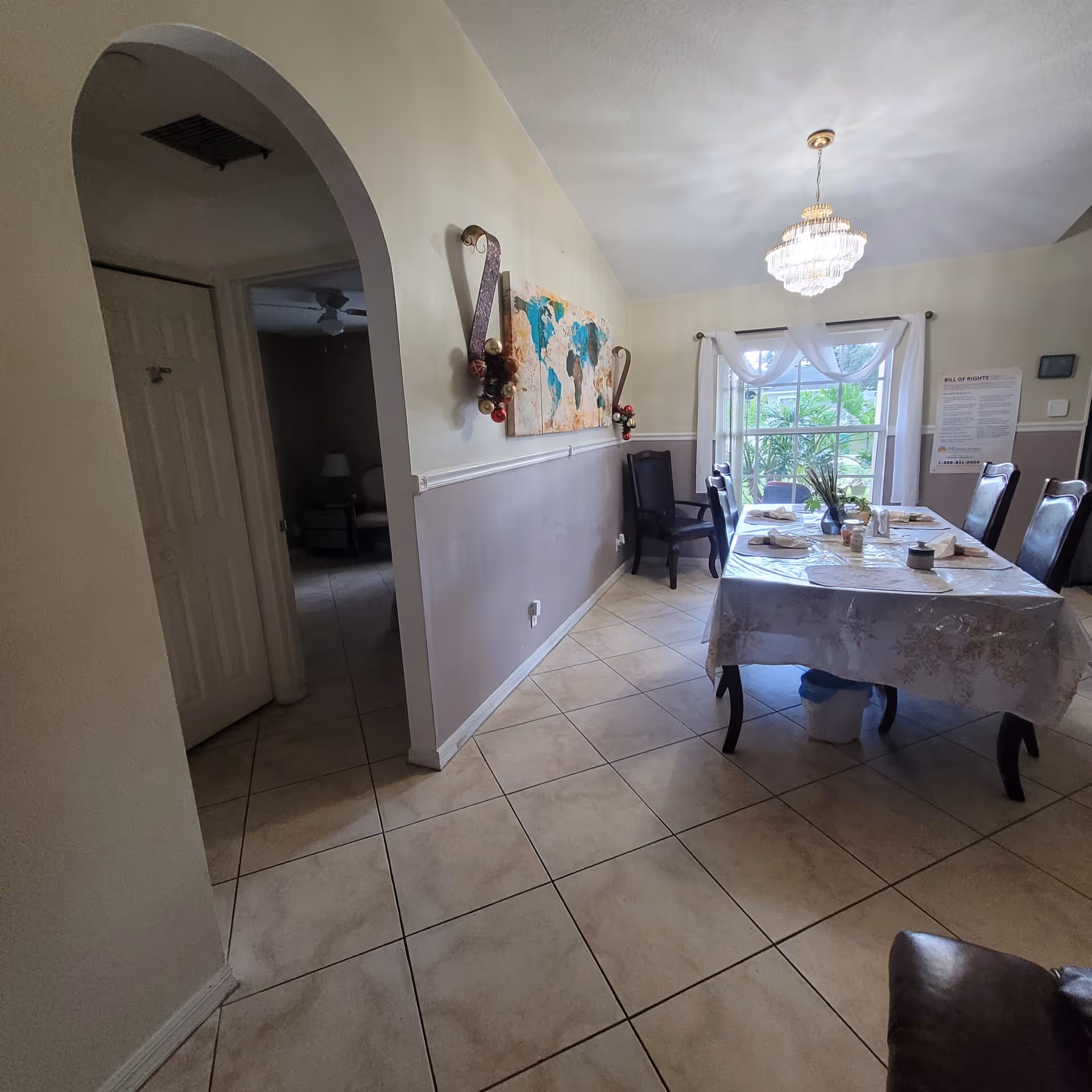 A dining room with a rectangular table covered with a floral tablecloth and set with napkins and condiments. There are six dark wooden chairs around the table. A large window with white curtains lets in natural light, and a chandelier hangs from the ceiling. On the wall, there is a world map artwork and decorative wall sconces with holiday ornaments. The floor is tiled, and an arched doorway leads to another room.