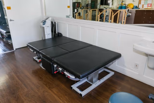A therapy or examination room with a black padded treatment table, a white sink mounted on the wall, a blue stool, and medical equipment including a scale and parallel bars reflected in a large mirror on the wall.