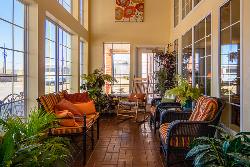 Sunlit indoor sitting area with wicker chairs, a rocking chair, cushions and many potted plants beside large windows.