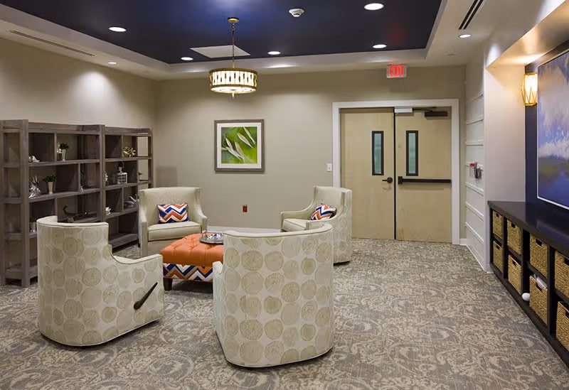 A cozy seating area in a senior living facility with four patterned armchairs arranged around an orange ottoman. The room features a patterned carpet, a wooden shelving unit with decorative items, a framed green leaf artwork on the wall, double beige doors, and a modern chandelier hanging from a recessed ceiling.
