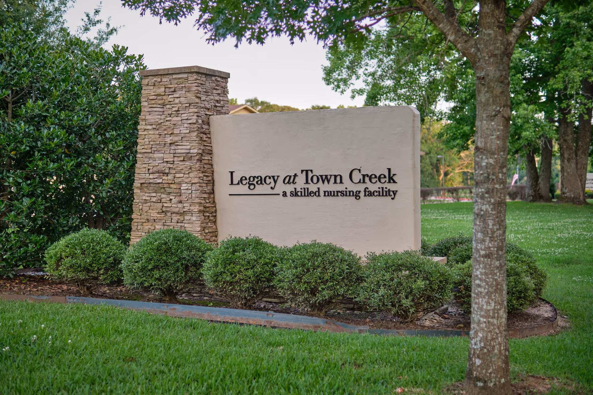Outdoor view of a sign for Legacy at Town Creek, a skilled nursing facility, surrounded by green bushes and trees with a stone pillar on the left side.