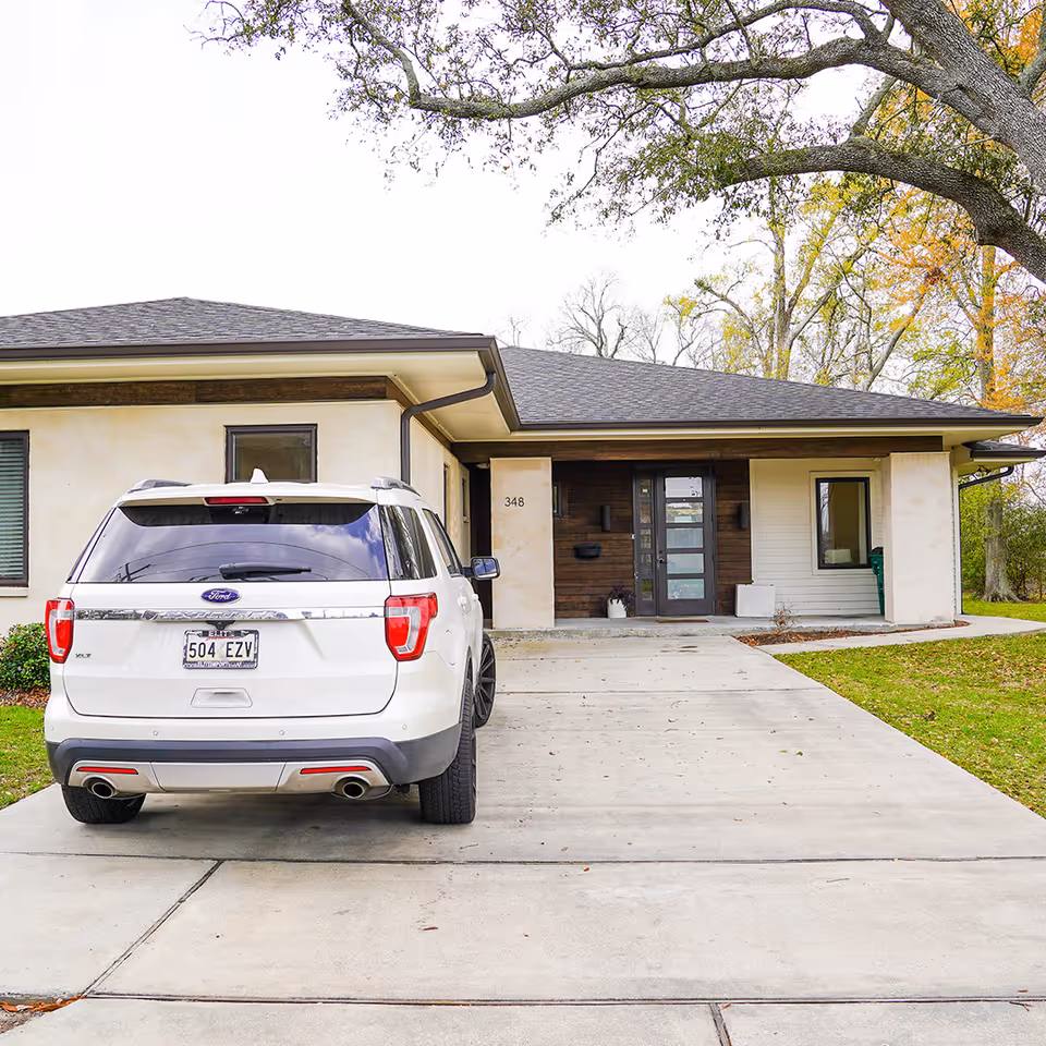 Front exterior view of a single-story residence with a modern design, featuring a white Ford SUV parked in the driveway. The house has a combination of light-colored stucco and dark wood paneling around the entrance, with a glass front door and a small porch area. Trees with autumn foliage are visible in the background.