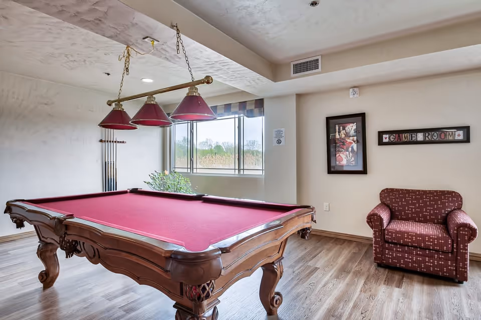 Interior view of a game room featuring a red felt pool table with wooden legs, a matching red armchair, a window with a view of greenery outside, a wall-mounted rack holding pool cues, a framed picture, and a sign that reads 'GAME ROOM'.