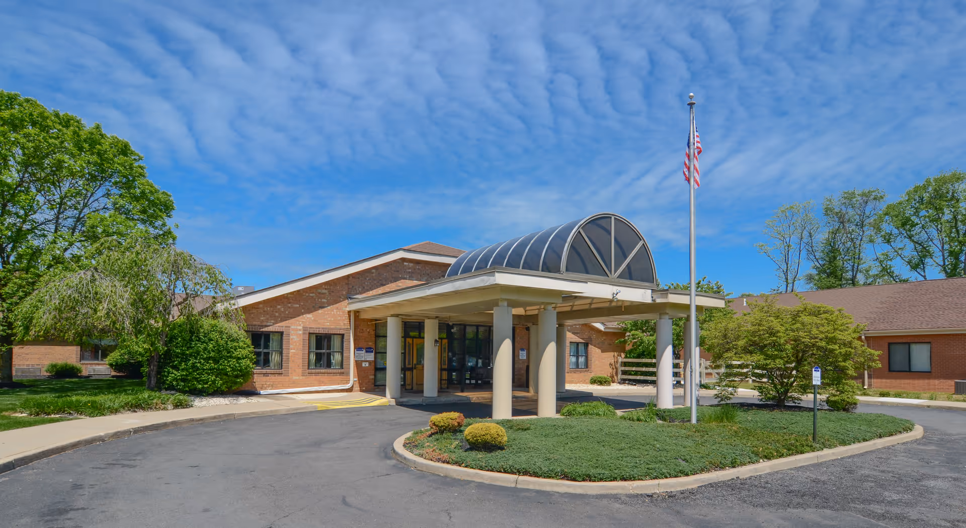 Exterior view of Yardley Rehabilitation and Healthcare Center showing the main entrance with a covered driveway, an American flag on a flagpole, surrounding greenery, and a partly cloudy blue sky.