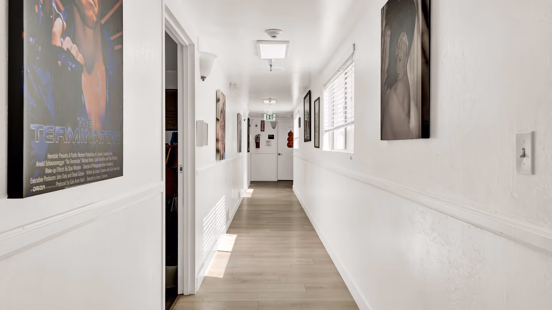 A bright, narrow hallway with light wooden flooring and white walls adorned with framed pictures and posters, including a visible poster of the movie The Terminator. There is a window with blinds on the right side and an exit door at the end of the hallway with an exit sign above it.