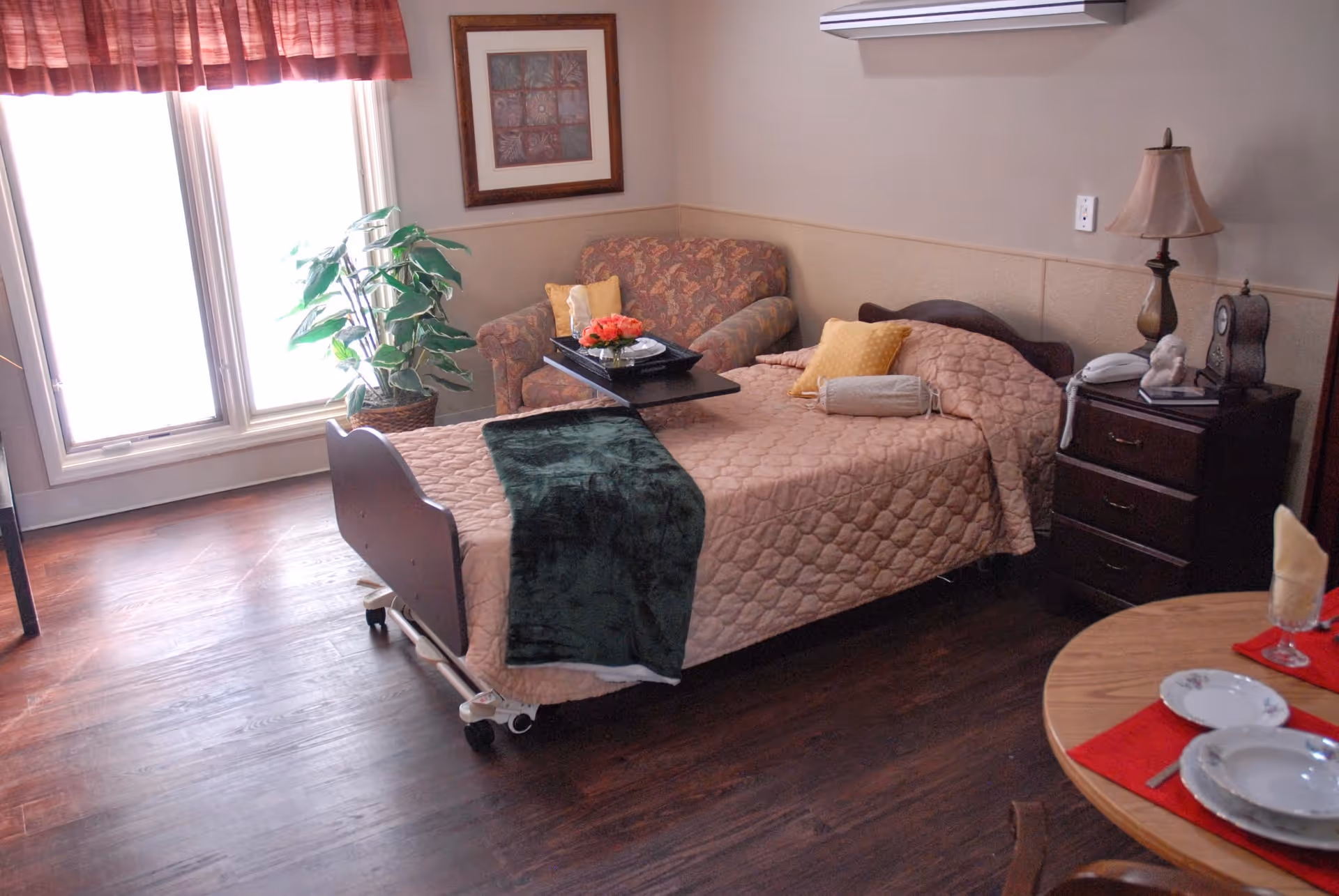 A cozy room with a single bed covered in a quilted pink bedspread and a dark green throw blanket. Next to the bed is a wooden nightstand with a lamp, a clock, and a telephone. Behind the bed is a floral-patterned armchair with a tray holding a flower arrangement and a cup. A large window with red curtains lets in natural light, and a potted plant is placed near the window. A round wooden table set with plates, glasses, and napkins is partially visible in the foreground.
