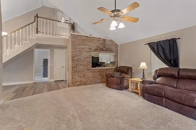 A spacious living room with beige carpet, two brown leather recliners, a wooden side table with a lamp, a ceiling fan with lights, and a brick wall with a pass-through window to the kitchen. There is a staircase with white railing leading to an upper floor, and a doorway leading to another room with a refrigerator visible.