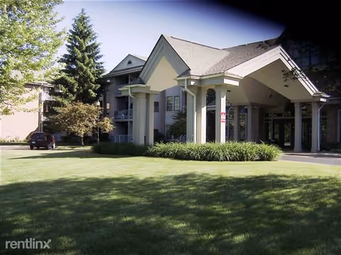Exterior view of a senior living facility building with a covered entrance supported by columns, surrounded by green grass and trees under a clear sky.