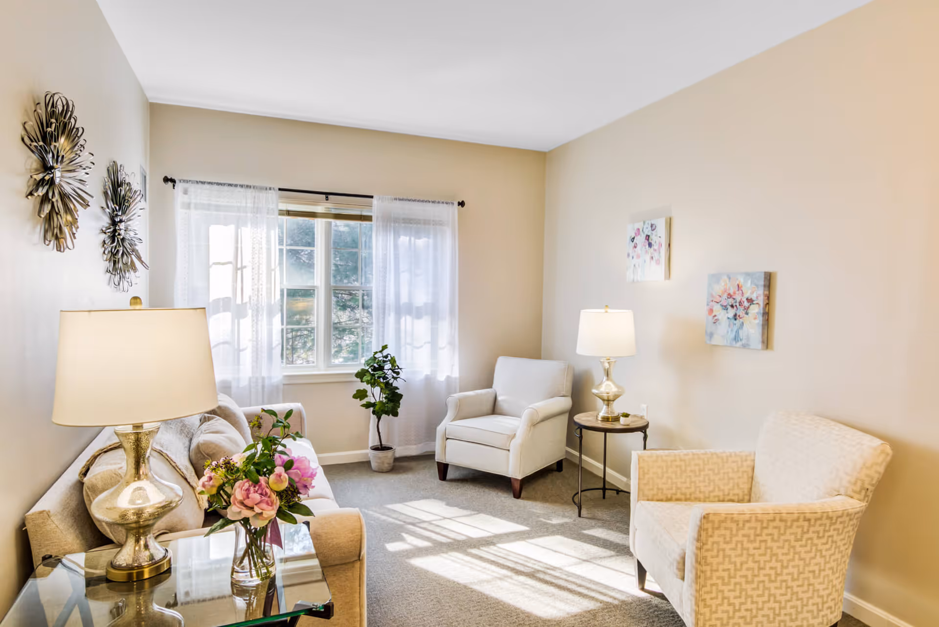 A bright and cozy living room with beige walls and carpet. The room features a beige sofa with cushions, two armchairs, a glass side table with a lamp and a vase of flowers, and a small round table with another lamp. There are decorative wall hangings and two floral paintings on the walls. A window with sheer white curtains lets in natural light, and a potted plant is placed near the window.