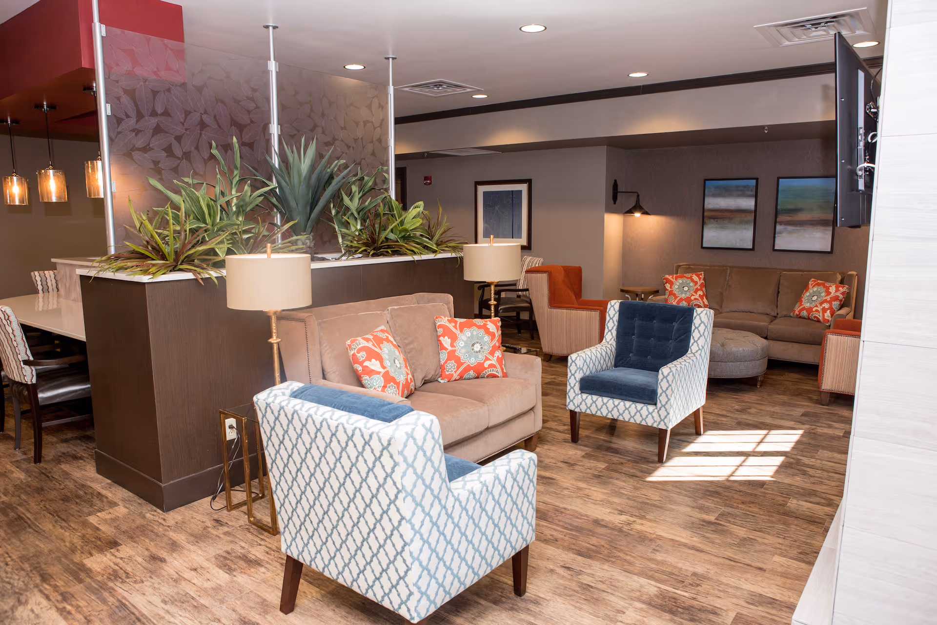 Communal living room with sofas, patterned armchairs, lamps, and decorative plants around a seating area.