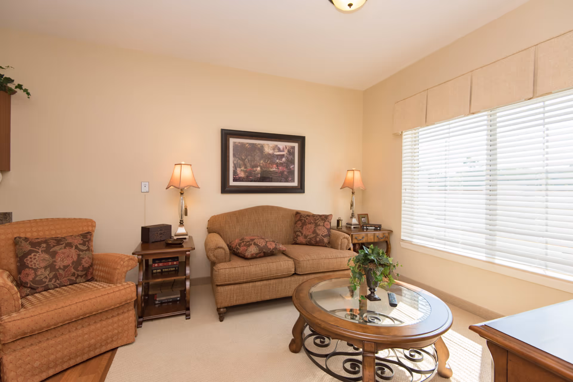 A cozy living room with a beige loveseat and an orange armchair, both adorned with floral-patterned cushions. Between them are two wooden side tables with lamps and decorative items. A round glass coffee table with a wooden frame and a potted plant sits in front of the loveseat. Large window blinds allow natural light to fill the room, and a framed picture hangs on the wall above the loveseat.