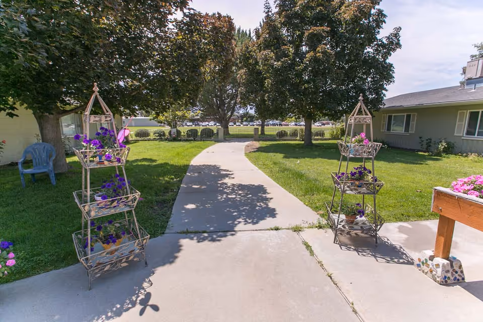Outdoor pathway in a garden area of a senior living facility with green grass, trees, and decorative metal plant stands holding purple flowers on either side of the concrete path. A building with windows and a roof is visible on the right side.