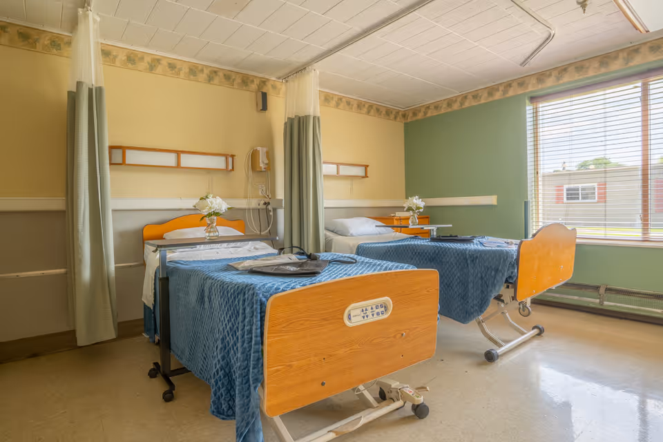 A nursing facility room with two hospital beds covered with blue blankets, each with a small table and a vase of white flowers. The room has light yellow and green walls, a large window with blinds, and privacy curtains between the beds.