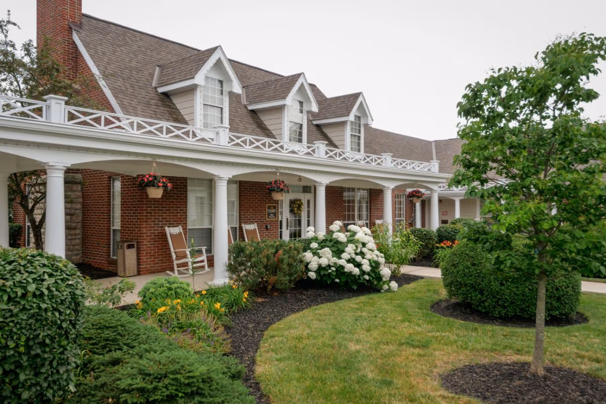 Exterior view of a brick building with a covered porch featuring white columns and rocking chairs. The porch is decorated with hanging flower baskets and surrounded by well-maintained landscaping including bushes, flowering plants, and a small tree.