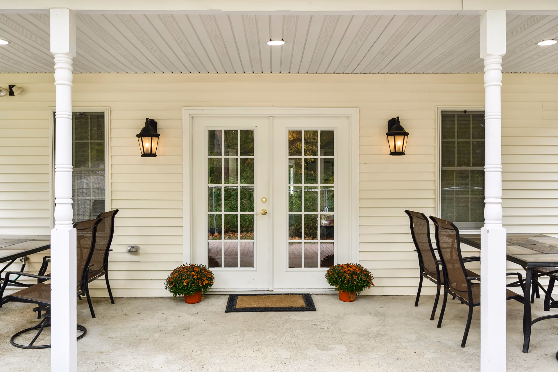 Covered outdoor patio area with white pillars and a white ceiling. There are two glass-paneled double doors in the center with two black wall-mounted lantern lights on either side. Two potted plants with orange flowers are placed symmetrically on the ground near the doors. On both sides of the patio, there are black metal chairs and tables.