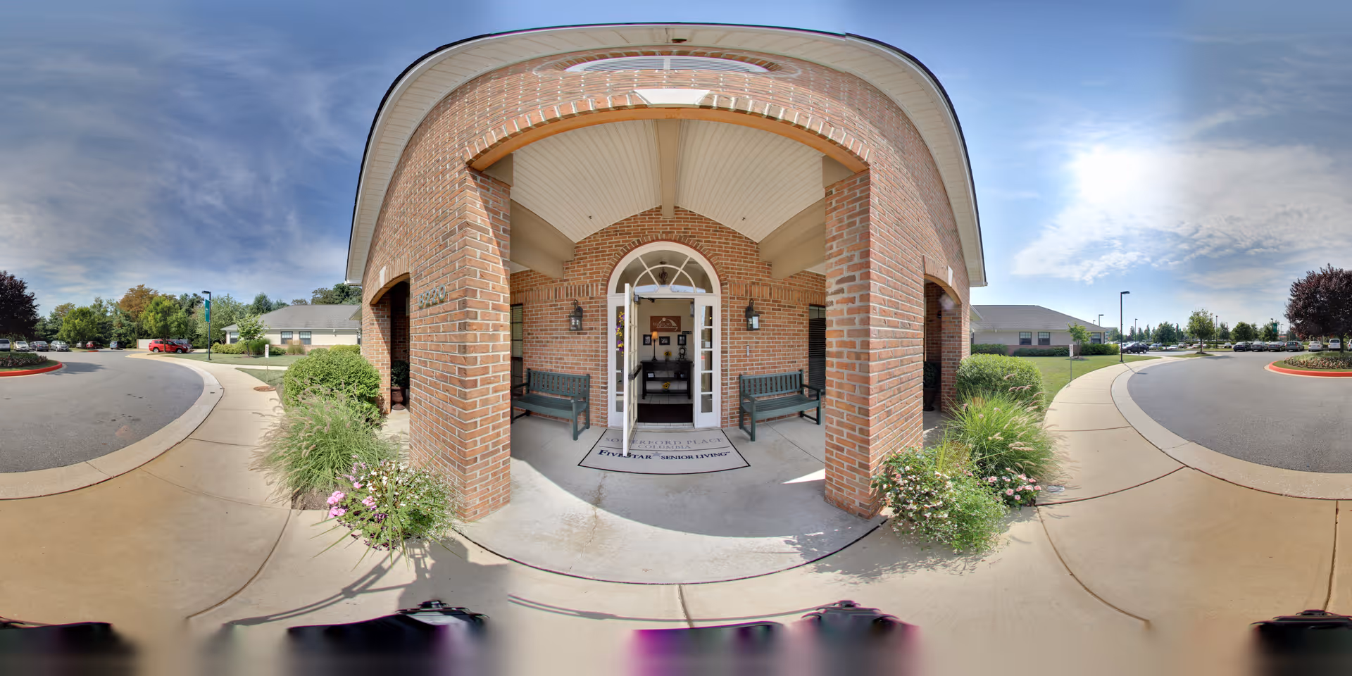 Front entrance of a brick building with a covered porch supported by brick columns. Two green benches are placed on either side of the entrance door. There are plants and flowers along the walkway leading to the entrance. The sky is partly cloudy with sunlight illuminating the scene.