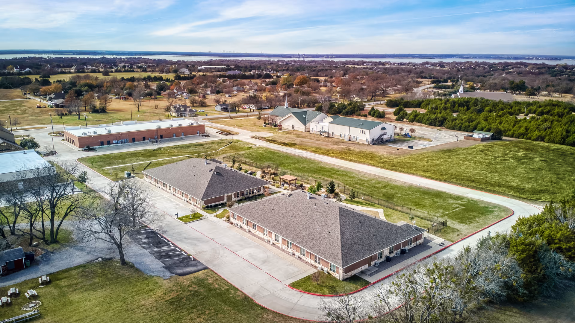 Aerial view of AvilaCare Assisted Living and Memory Care of Heath facility showing multiple single-story buildings with pitched roofs, surrounded by green lawns, trees, and paved driveways. The landscape includes nearby residential areas and open fields under a partly cloudy sky.