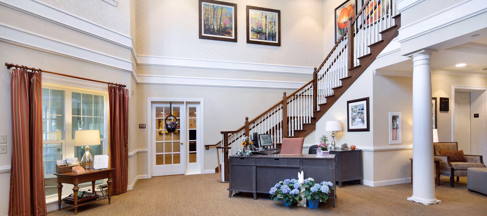 Reception lobby of a senior living facility with a front desk, staircase, seating area, and artwork on the walls.