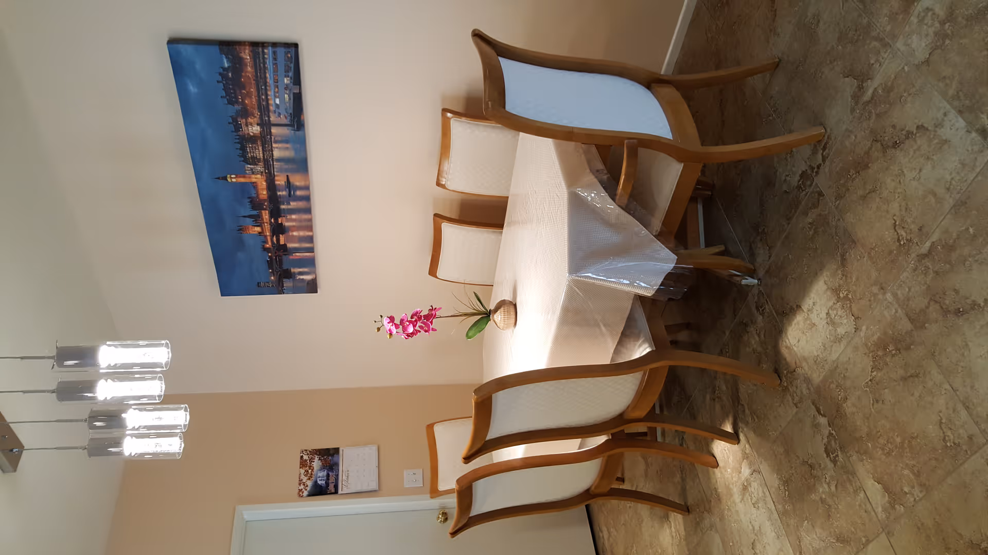 A dining room with a rectangular table covered with a clear plastic tablecloth and surrounded by six wooden chairs with white cushioned backs and seats. A small vase with a pink orchid is placed in the center of the table. Above the table hangs a modern light fixture with four cylindrical lights. On the wall behind the table is a large framed picture of a cityscape at night, featuring illuminated buildings and a river. A calendar is visible on the adjacent wall near a closed door.