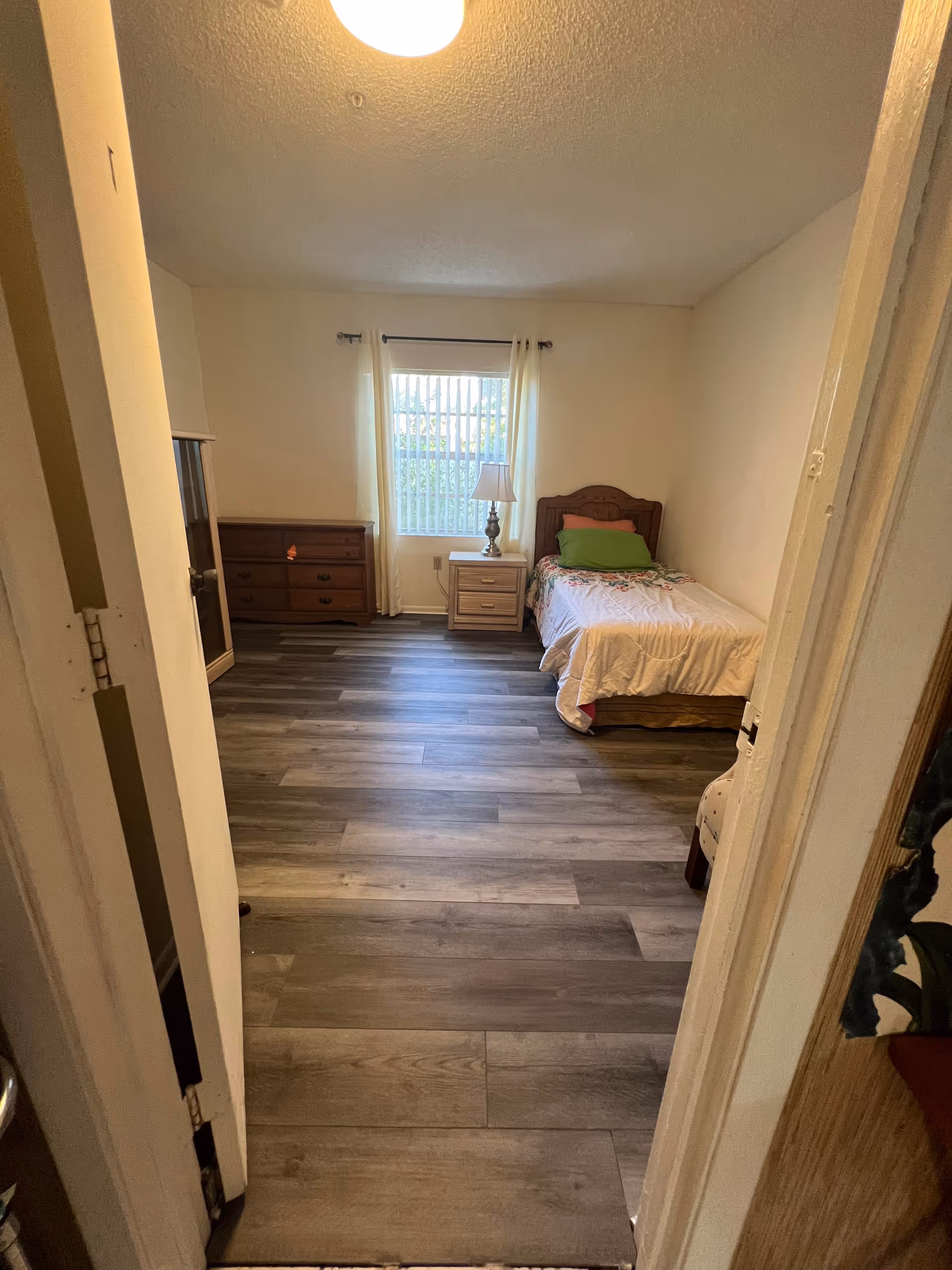 View into a bedroom with a single bed on the right side covered with a white blanket and green pillow. There is a wooden nightstand with a lamp next to the bed, and a window with white curtains behind it. On the left side of the room, there is a wooden dresser and a cabinet. The floor has wood-like tiles and the walls are painted light beige.