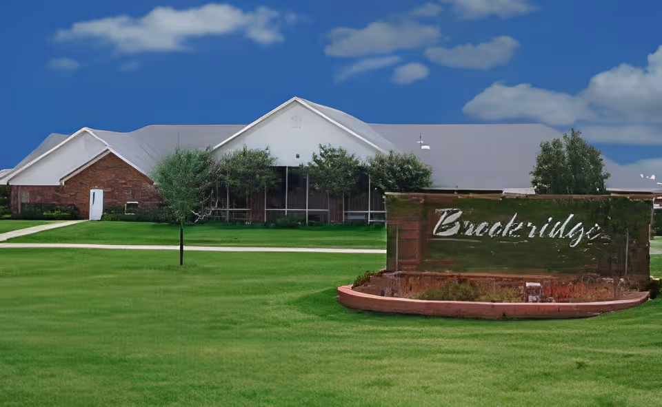 Front lawn and entrance sign reading 'Brookridge' with a single-story brick and white-roof retirement community building in the background under a blue sky.