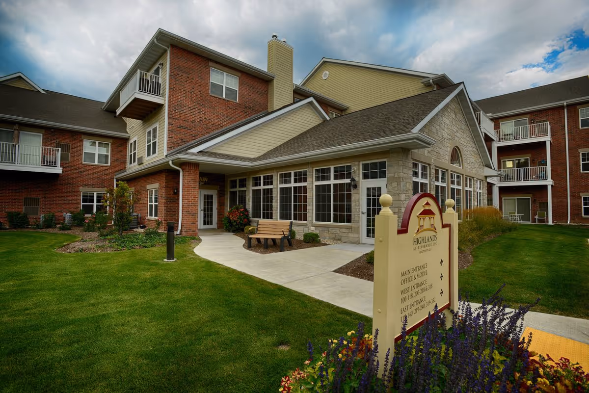 Exterior view of Highlands at Riverwalk Apartments 55+, showing a multi-story brick and stone building with large windows, a bench near the entrance, well-maintained green lawn, and a sign with directions to various entrances.