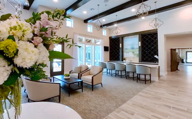 A bright and spacious living room area with high ceilings and large windows. The room features a seating area with beige armchairs and a coffee table, a bar counter with four bar stools, and decorative pendant lights hanging from the ceiling. There is a large floral arrangement in the foreground and a large painting or screen on the wall behind the bar. The flooring is a combination of carpet and tile.