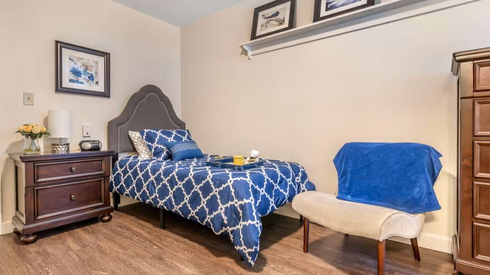 A bedroom with a single bed featuring a blue and white patterned comforter and multiple pillows. Next to the bed is a dark wooden nightstand with a lamp, a small radio, and a vase of flowers. There is a beige cushioned chair with a blue blanket draped over it. The walls are light-colored with framed artwork and a shelf holding decorative items. The floor is wooden.