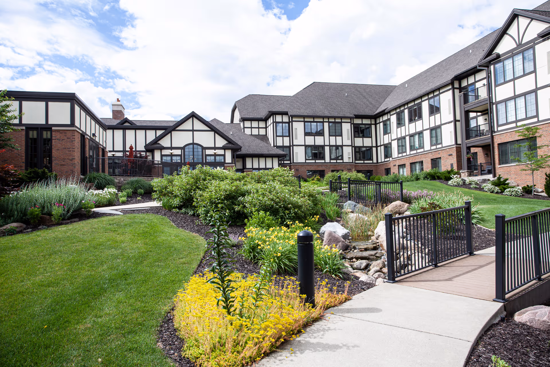 A landscaped outdoor area of a senior living facility with green grass, flowering plants, shrubs, a small bridge over a rock-lined stream, and a multi-story building with Tudor-style architecture in the background under a partly cloudy sky.
