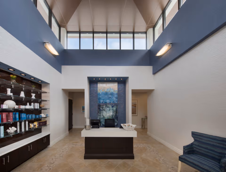 Interior view of a modern reception area with a high ceiling and large windows near the ceiling. The space features a central desk with two chairs behind it, a decorative blue and white wall art piece, and a display shelf on the left side with various products. There is a blue striped armchair on the right side and beige tiled flooring.