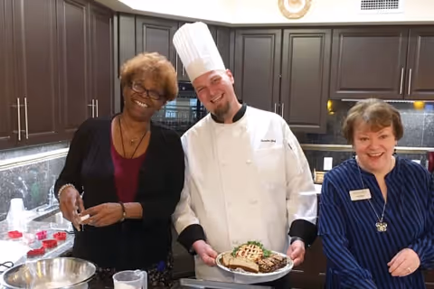 Three smiling staff members, including a chef in a tall white hat holding a plated dish, stand together in a residential-style kitchen.