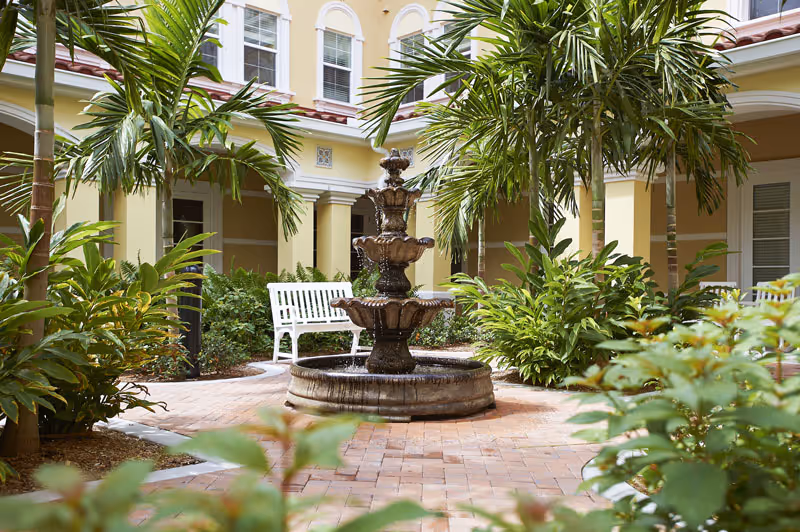 A sunny courtyard with a three-tiered stone fountain surrounded by tropical plants, a white bench, and a yellow building façade.