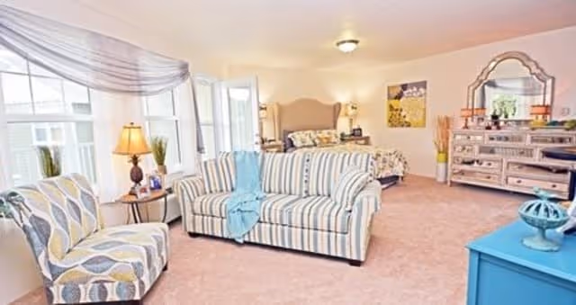 A bright and spacious bedroom with a large bed in the background, a striped sofa and patterned armchair in the foreground, a blue throw draped over the sofa, a dresser with a large mirror, and a window with sheer curtains letting in natural light.