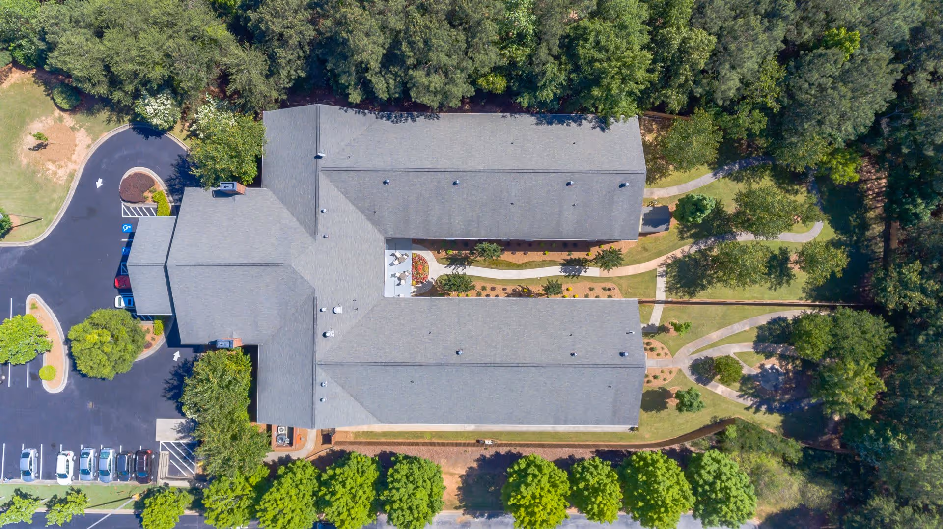 Aerial view of a senior living facility building with a gray roof surrounded by trees and landscaped gardens. There are paved walkways around the building and a parking lot with several cars on the left side.