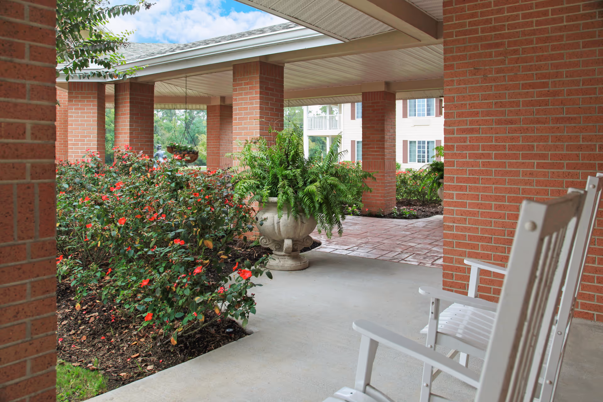 Covered outdoor patio area with brick columns and a concrete floor. There are white rocking chairs on the right side and a large planter with green ferns in the center. Flowering bushes with red blooms are on the left side, and a building with windows is visible in the background.