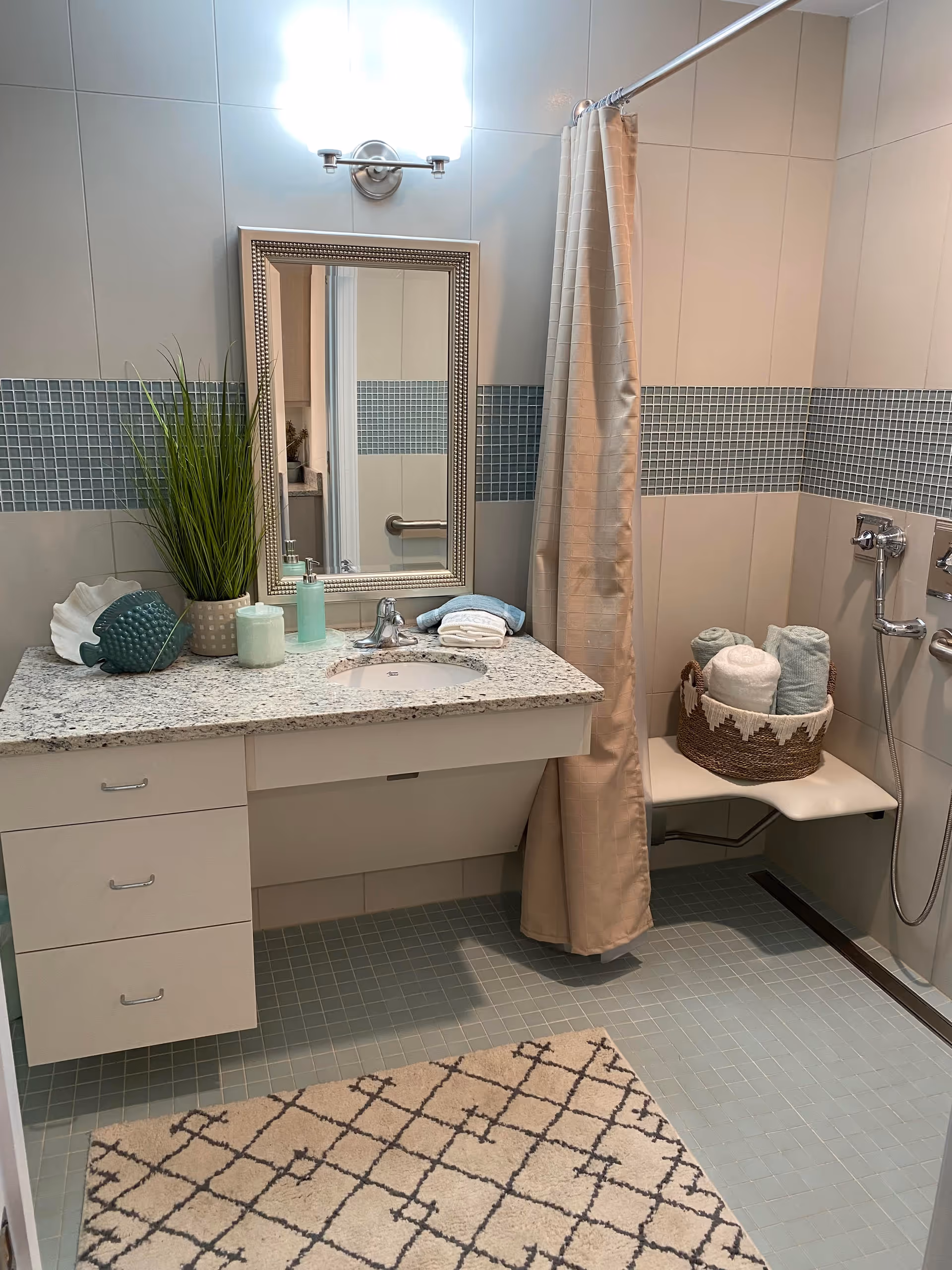 A clean and modern bathroom featuring a granite countertop with a sink, a decorative mirror with a silver frame, and a wall-mounted light above it. There is a beige shower curtain partially covering a shower area with a fold-down seat holding a basket filled with rolled towels. The walls have a blue mosaic tile accent strip, and the floor is tiled in a matching blue. A patterned beige and black rug is placed on the floor in front of the sink. Decorative items including a green plant, a ceramic fish, and soap dispensers are on the countertop.
