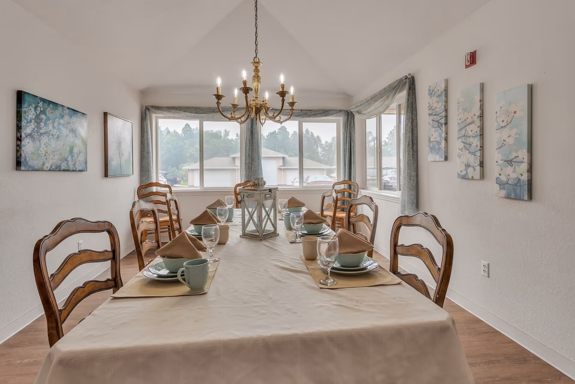 A dining room with a long table set for six people. The table is covered with a beige tablecloth and has place settings including plates, bowls, mugs, glasses, and brown napkins. A decorative lantern is placed in the center of the table. The room has wooden chairs, large windows with light blue curtains, and floral artwork on the walls. A chandelier with lit bulbs hangs above the table.