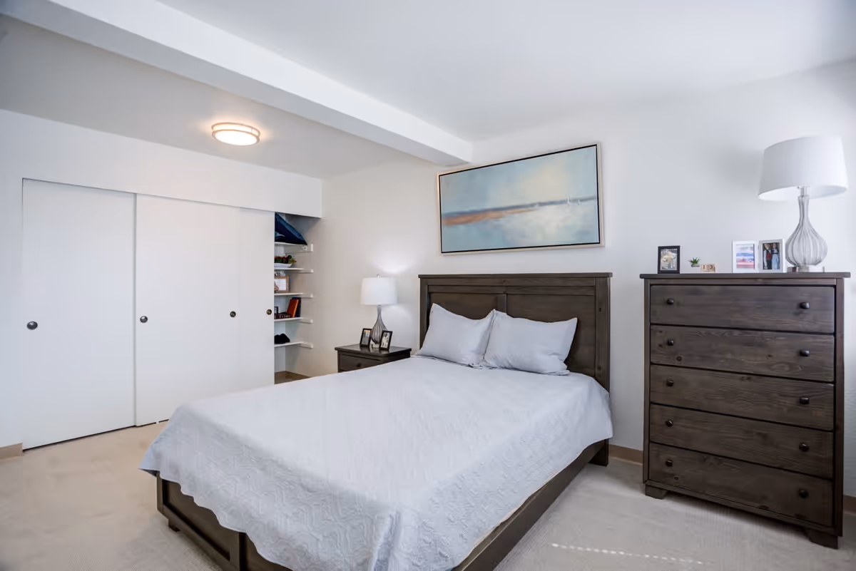 A bedroom with a wooden bed frame and white bedding, two pillows, a wooden nightstand with a lamp and picture frames, a wooden dresser with a lamp and framed photos, white walls, a large painting above the bed, and a closet with sliding white doors.