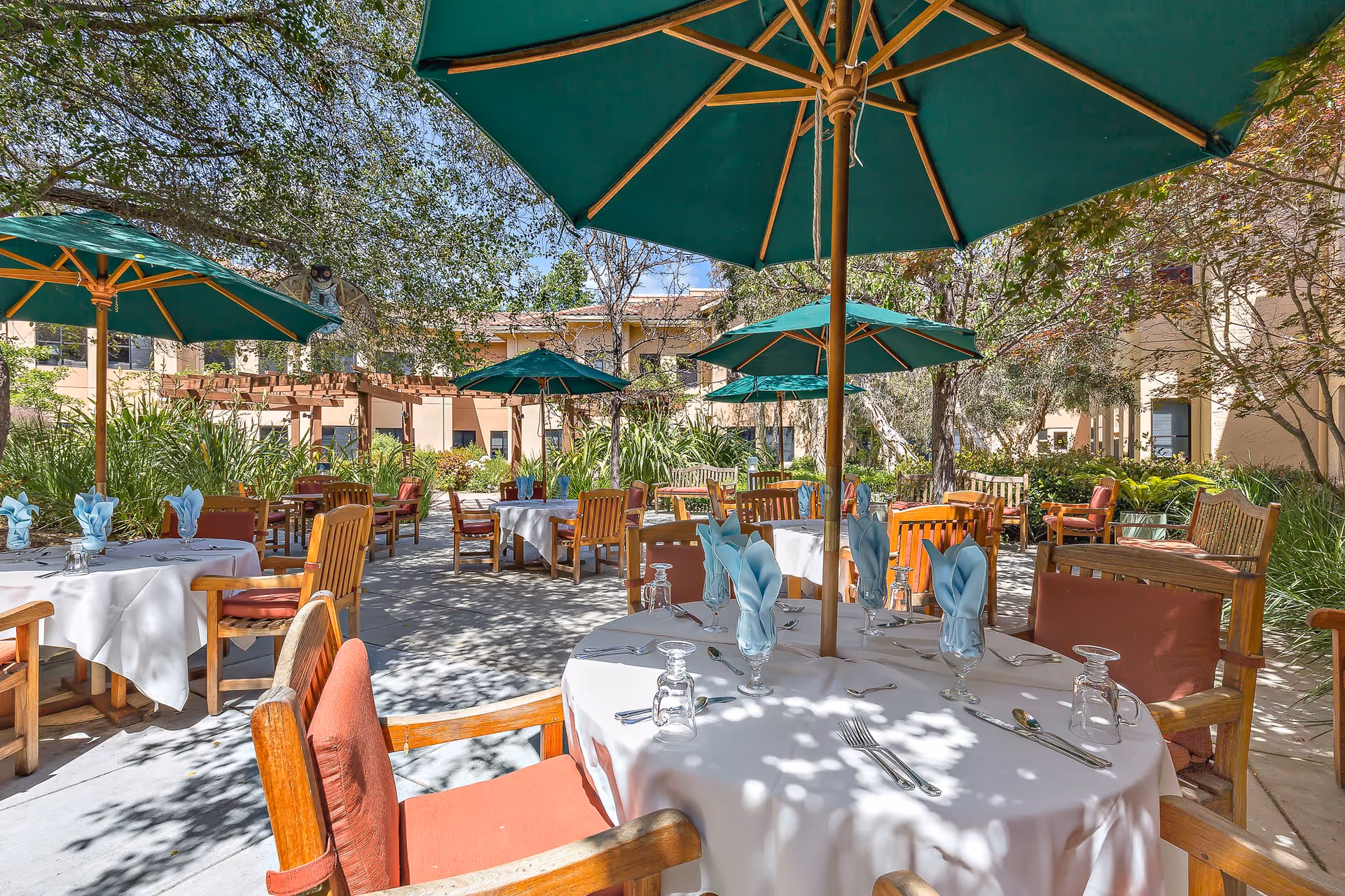 Outdoor dining area at Saratoga Retirement Community Assisted Living with round tables covered in white tablecloths, set with blue folded napkins, glasses, and silverware. Wooden chairs with red cushions surround the tables, and large green umbrellas provide shade. The area is surrounded by trees, plants, and a beige building in the background under a clear blue sky.