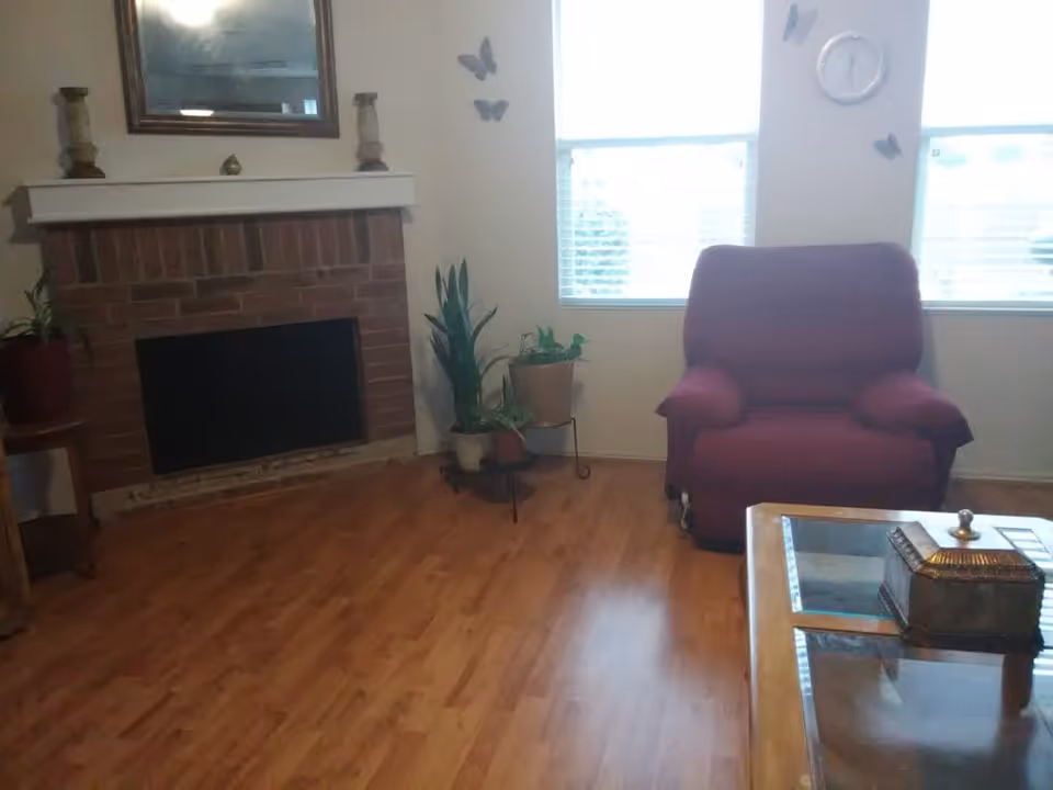 A cozy living room with a brick fireplace on the left, a large mirror above the mantel, and two candle holders. There are several potted plants near the fireplace and a large window with blinds letting in natural light. A comfortable maroon armchair is positioned near the window, and a wooden coffee table with a decorative box is in the foreground.