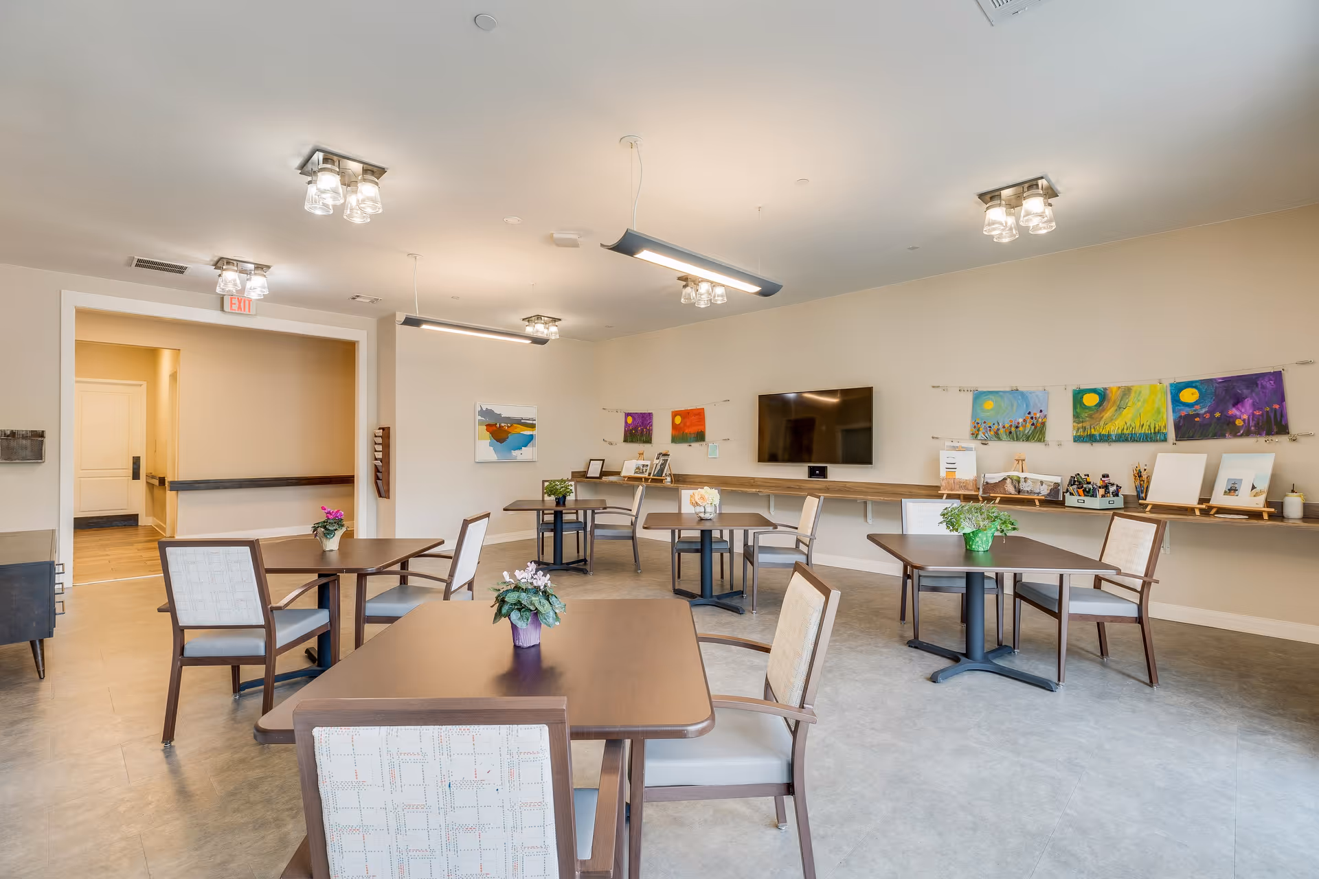 A bright, spacious common room with several square wooden tables and cushioned chairs arranged around the room. Each table has a small potted plant as decoration. The walls are light-colored and adorned with colorful paintings and artwork. A flat-screen TV is mounted on the far wall above a long wooden shelf that holds various items and art supplies. The room has modern ceiling lights and an exit door visible in the background.