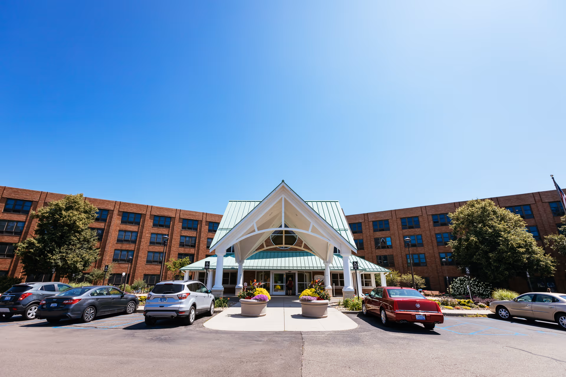 Front exterior view of Glacier Hills Senior Living Community building with a green roof entrance canopy, several parked cars, trees, and clear blue sky.