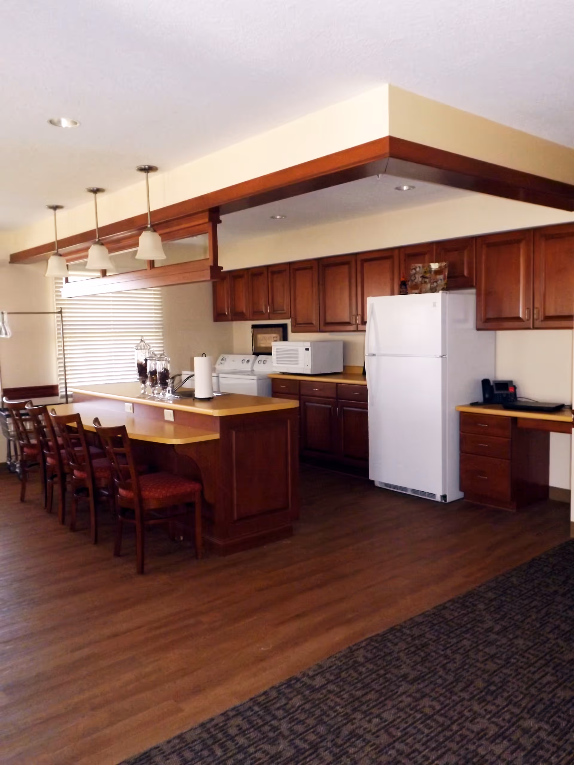 Interior view of a kitchen area with wooden cabinets, a white refrigerator, microwave, and a washing machine. There is a long counter with several wooden chairs with red cushions. Pendant lights hang from the ceiling above the counter. The floor is wooden and there is a carpeted area adjacent to the kitchen.