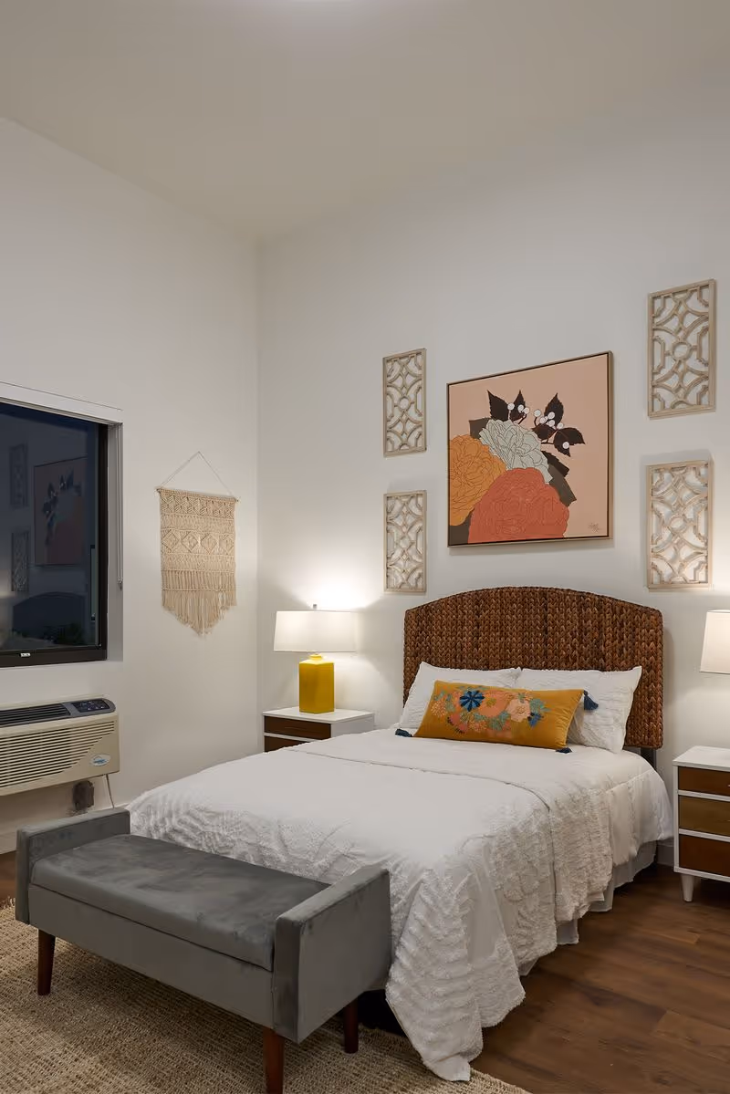 A cozy bedroom with a white bedspread, a woven headboard, and a decorative floral pillow. There are two bedside tables with lamps, wall art including a large floral painting and four decorative panels, a macrame wall hanging, a window with a view outside, and a gray upholstered bench at the foot of the bed.