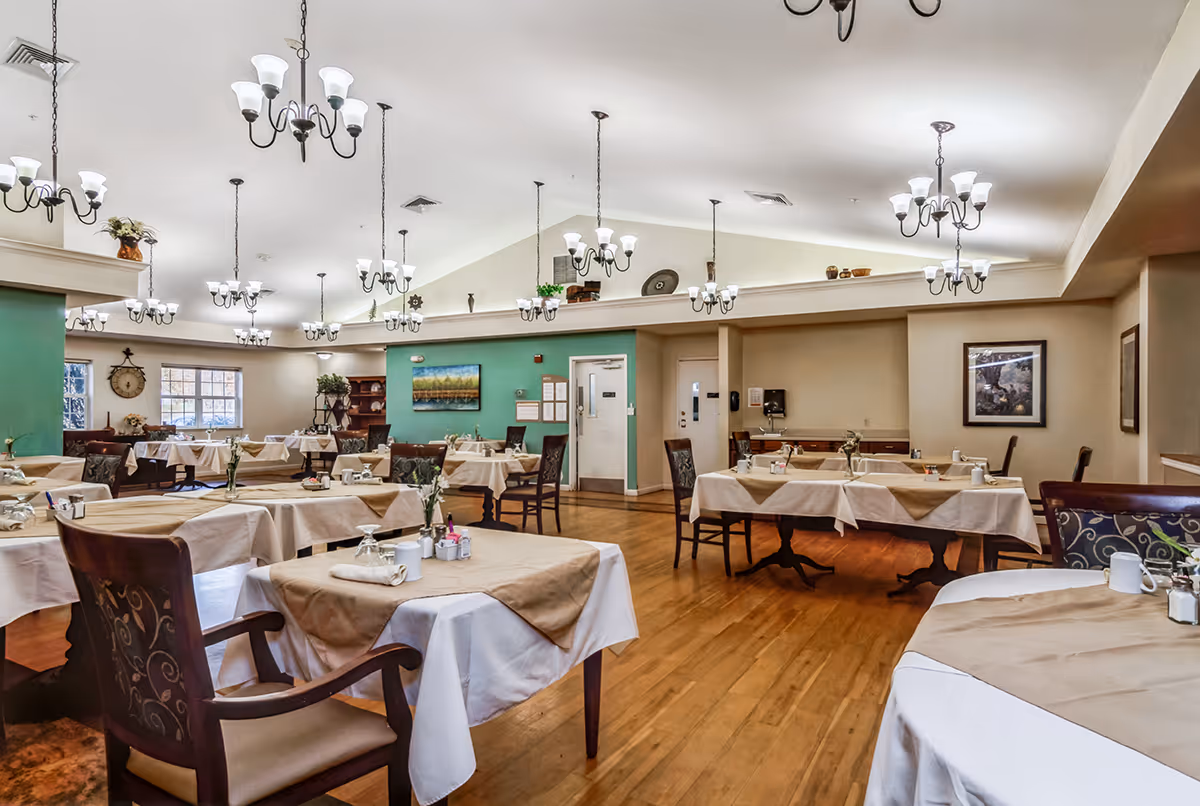A spacious dining room with multiple tables covered in white and beige tablecloths, each set with glasses, napkins, and small flower vases. The room has wooden floors, green and beige walls, several chandeliers hanging from the ceiling, and framed artwork on the walls.