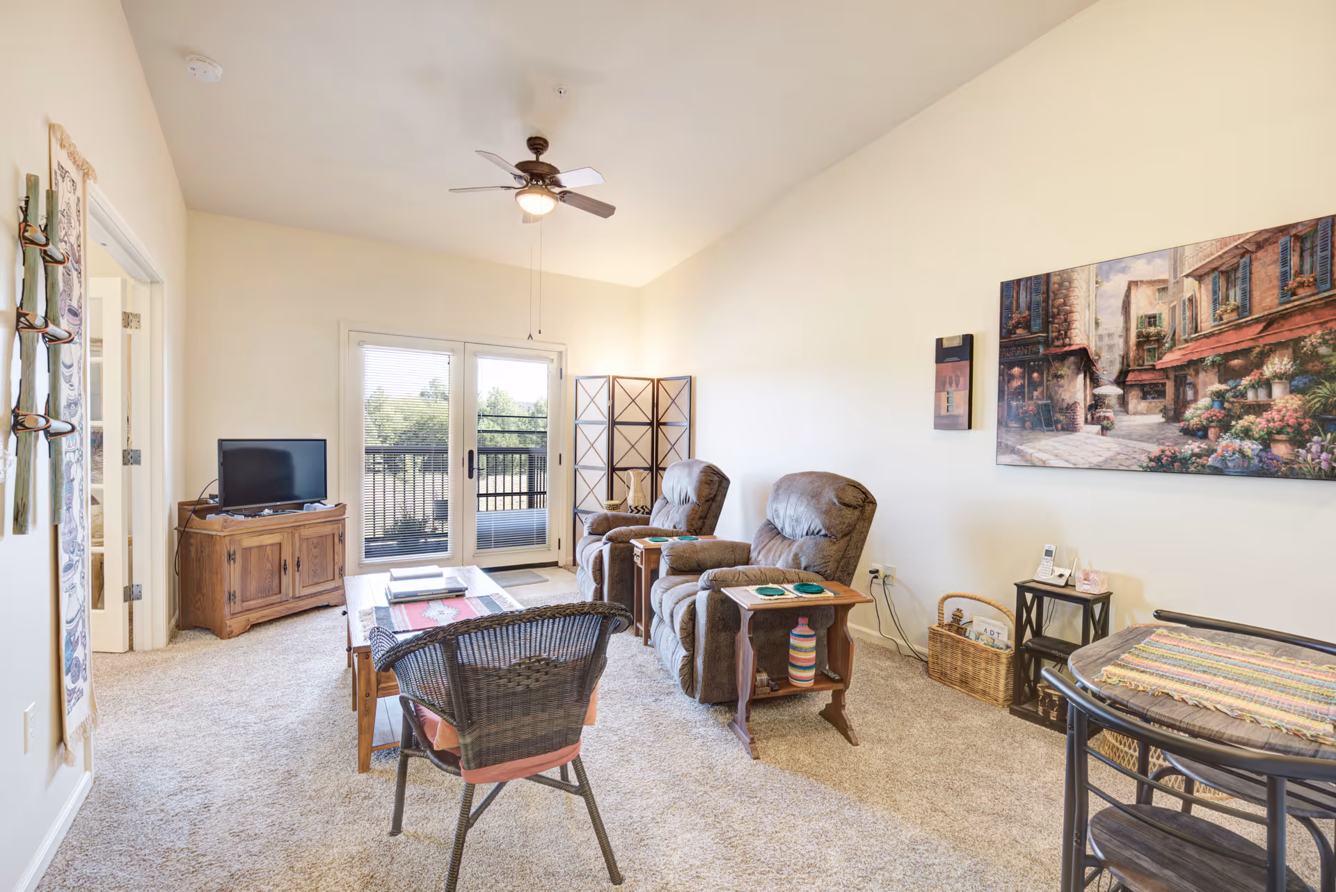 Bright living room with two recliner chairs, a wicker chair and coffee table facing a TV, and sliding doors opening to a balcony.